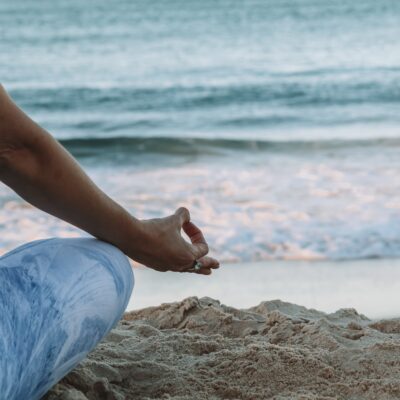 A person sits cross-legged on a beach, facing ocean waves in a meditative pose.