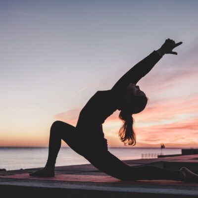 The profile of a woman practicing yoga is silhouetted against a waterfront sunset.
