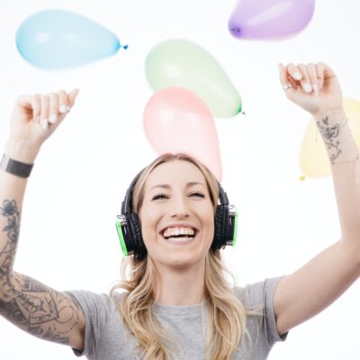 A woman wearing headphones lifts her arms in a joyful dance, while balloons float overhead.
