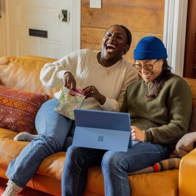 Two friends sit on a couch, laughing together and looking at a digital tablet.