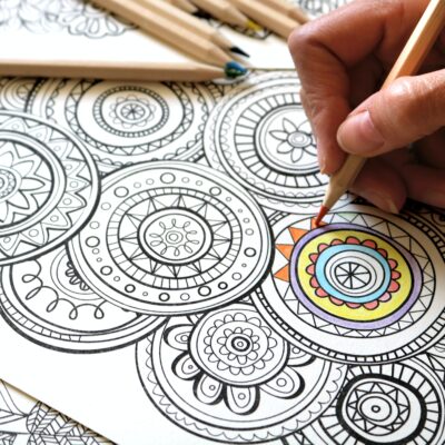 A hand grasps a colored pencil, coloring in a black and white mandala illustration